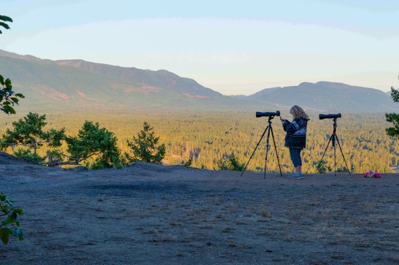 Silhouette of Sharron standing by her tripod with her camera mounted on it, with sunlit hills in the backgroundl