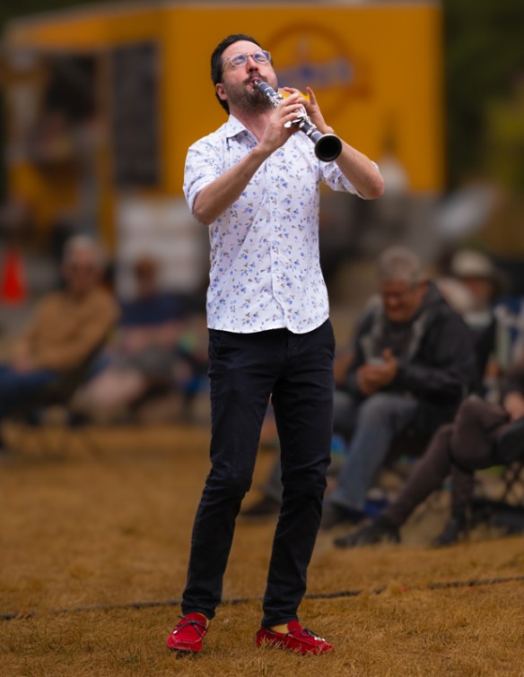 A man standing, playing the clarinet in front of an audience who are blurred in the background. The man's head is tilted back and his eyes are closed.